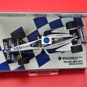 Op de afbeelding: Een model van de Williams BMW FW22 Formule 1-auto, gepresenteerd in een transparante display case. De auto is wit en marineblauw met sponsorlogo's. Het model staat op een blauw-witte geblokte achtergrond.