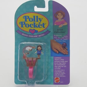 May include: Polly Pocket Rose Dream Ring Jewel Collection toy. The packaging is teal and purple, with a clear plastic window showing a miniature doll and a pink and gold ring. The ring is designed to hold the doll.