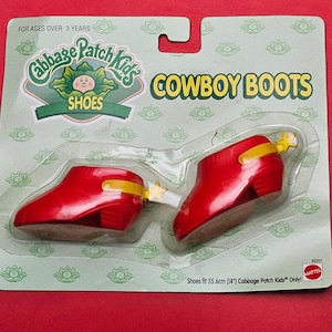 May include: Red plastic Cabbage Patch Kids cowboy boots in a clear plastic package. The boots have yellow accents and a star-shaped detail. The packaging reads "COWBOY BOOTS" and "SHOES". The boots fit 35.6cm dolls.