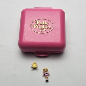 Op de afbeelding: Een roze Polly Pocket compact met een goudkleurig ovaal logo en hart. Inclusief een kleine gele berenfiguur en een miniatuurpop met blond haar en een roze jurk.