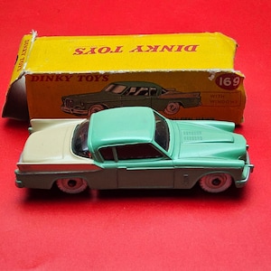 May include: A vintage Dinky Toys model car, a mint green and cream-coloured coupe, sits in front of its yellow box. The box features an illustration of a similar car. The toy car has white-walled tyres and is set against a red background.
