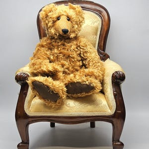 Puede incluir: Un oso de peluche marrón dorado con pelaje texturizado está sentado en una silla de madera antigua. La silla tiene un asiento y respaldo de tela crema estampada, con brazos y patas de madera marrón oscuro. El oso tiene una nariz negra y patas marrones.
