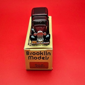 Könnte beinhalten: Ein schwarzes Oldtimer-Modellauto steht auf seiner Originalverpackung vor rotem Hintergrund. Das Auto hat einen detaillierten Chromgrill und Scheinwerfer. Die Box ist beige und trägt die Aufschrift "Brooklin Models".