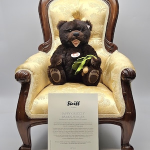 May include: A dark brown teddy bear with a light brown snout and paws, seated in a vintage gold upholstered chair with dark wood trim. The bear holds a green and brown leaf-like object. A white card with text is in front of the chair.