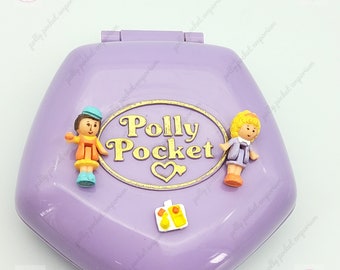 May include: Purple Polly Pocket toy with a gold heart and the words "Polly Pocket" on the front. The toy features two small figures, a boy and a girl, and a small yellow and orange object.