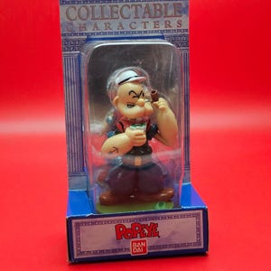 May include: A Popeye collectible figure in a clear plastic case, packaged in a blue and white box. The figure is of Popeye the Sailor Man, holding a pipe and wearing his signature outfit. The box reads "COLLECTABLE CHARACTERS" and "Popeye".