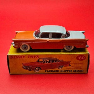 Op de afbeelding: Een vintage Dinky Toys Packard Clipper Sedan speelgoedauto, oranje en wit, staat op zijn originele gele doos. De doos heeft een rode illustratie van de auto en de tekst "Packard Clipper Sedan".