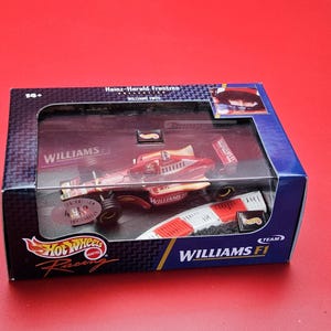Op de afbeelding: Een Hot Wheels Williams F1 raceauto model in een displaydoos. De auto is rood en goudkleurig met zwarte wielen. De doos is zwart en blauw met het Hot Wheels Racing logo en Williams F1 branding.
