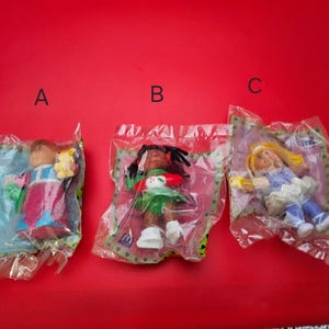 Peut inclure: Trois poupées vintage dans un emballage plastique, chacune avec des tenues et des coiffures différentes. Les poupées sont sur un fond rouge. Une poupée a les cheveux blonds, une autre a les cheveux foncés et la troisième a les cheveux châtain clair.