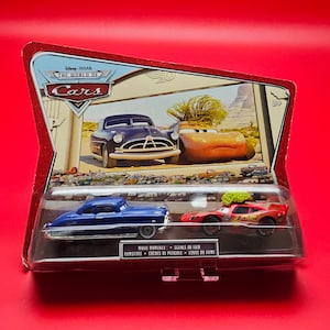 May include: A packaged set of two toy cars from Disney Pixar's "Cars" film. The set includes a blue car and a red race car, displayed in a clear plastic case with a themed backdrop. The packaging features the "World of Cars" logo.