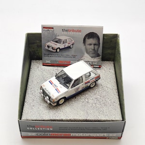Op de afbeelding: Een wit en blauw model van een Vauxhall Nova Sport 1300cc rallyauto, gepresenteerd in een doos. Het modelauto heeft Shell Oils-branding en het nummer 85. De verpakking bevat een foto van Colin McRae en de tekst "the tribute collection."