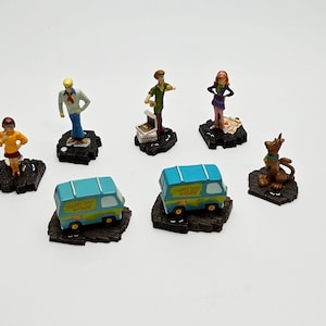 Pode incluir: Uma coleção de figuras em miniatura com personagens e veículos da série animada. As figuras incluem uma carrinha, um cão e vários personagens humanos, todos em bases castanhas escuras. A carrinha é azul clara com letras amarelas.