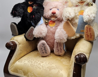 May include: Three plush teddy bears in black, pink, and white sit on a gold patterned chair. Each bear has a bell collar and a tag. The black bear has blue eyes, the pink bear has tan paws, and the white bear has a red nose.