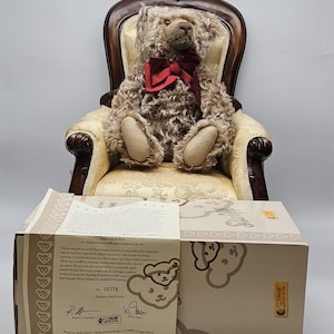May include: A plush teddy bear with curly brown fur and a red bow tie sits in a vintage armchair. The bear is positioned above a beige box with text and illustrations. The box has a label with the number 01771.