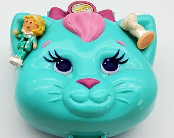 Può includere: Un giocattolo di plastica blu turchese a forma di gatto con un fiocco rosa, un naso rosa e ciglia nere. Il giocattolo ha una piccola apertura sul fondo e include una piccola bambola e un accessorio bianco e rosa.