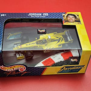 Op de afbeelding: Een boxed Hot Wheels Racing Jordan 199 modelauto. De gele raceauto heeft zwarte accenten en het nummer 8. De doos bevat een foto van een coureur en het Hot Wheels logo.