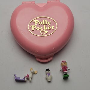 Può includere: Un compatto Polly Pocket rosa a forma di cuore con scritte dorate e un logo a forma di cuore. Include figurine in miniatura di un pupazzo di neve, una ragazza in abito rosa e una renna che tira una slitta viola. Un giocattolo vintage.