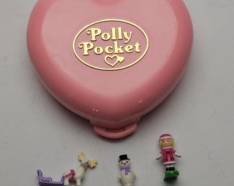 Könnte beinhalten: Ein rosa herzförmiges Polly Pocket Compact mit goldenen Buchstaben und einem Herzlogo. Enthält Miniaturfiguren eines Schneemanns, eines Mädchens in rosa Outfit und ein Rentier, das einen lila Schlitten zieht. Ein Vintage-Spielzeug.
