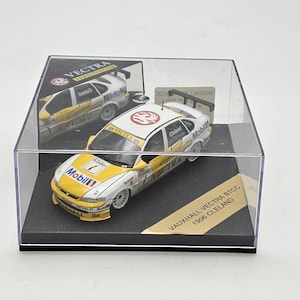 Op de afbeelding: Een model van een Vauxhall Vectra BTCC 1996 Cleland in een transparante vitrine. De auto is wit en geel met Mobil-branding en nummer 7. De vitrine staat op een zwarte basis.