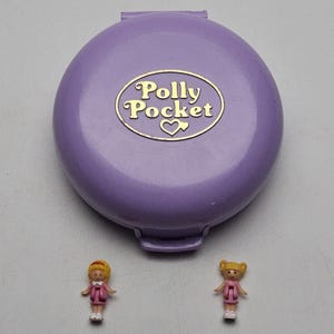 Puede incluir: Un compacto lila de Polly Pocket con un logotipo dorado y dos muñecas en miniatura. El compacto está cerrado y es ovalado. Las muñecas visten vestidos rosas y zapatos blancos.