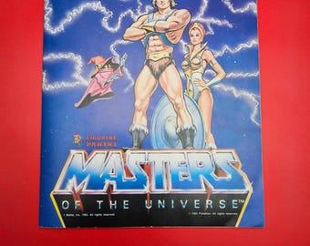 Puede incluir: Una portada de álbum de pegatinas Panini "Masters of the Universe" vintage. La portada presenta ilustraciones de He-Man, un personaje femenino y un mago sobre un fondo azul con rayos. El título "Masters of the Universe" se muestra de forma destacada.