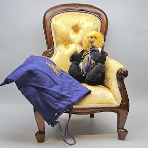 Könnte beinhalten: Ein Teddybär mit gelbem Kopf und schwarzem Körper, der eine lila Krawatte trägt, sitzt auf einem goldfarbenen, gepolsterten Stuhl. Ein blauer Kordelzugbeutel mit goldener Stickerei liegt auf dem Stuhl.