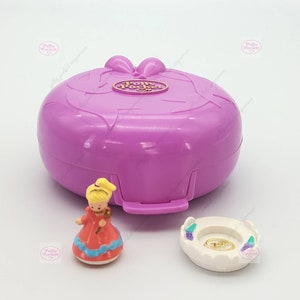 Könnte beinhalten: Ein lilafarbenes Polly Pocket Spielzeug mit einer goldenen Schleife und dem Schriftzug "Polly Pocket" auf der Vorderseite. Das Spielzeug ist geöffnet und zeigt eine kleine Plastikpuppe, die ein rotes Kleid und eine weiße Krone trägt. Außerdem gibt es einen kleinen weißen Plastik-Ringhalter mit einem goldenen Ring darin.