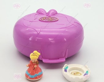 May include: A purple Polly Pocket toy with a gold bow and the words "Polly Pocket" on the front. The toy is open and shows a small plastic doll wearing a red dress and a white crown. There is also a small white plastic ring holder with a gold ring inside.
