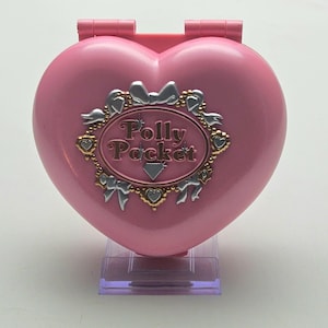 Op de afbeelding: Een roze hartvormige Polly Pocket compact met de woorden "Polly Pocket" in het midden. De compact heeft een zilveren en gouden decoratieve rand met strikken en harten. De compact staat op een doorzichtige plastic standaard.