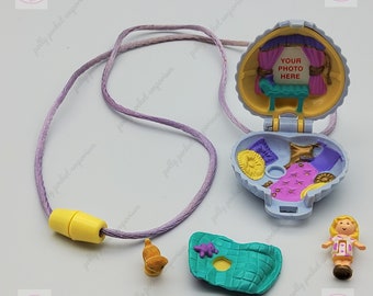 Könnte beinhalten: Eine lila und gelbe Polly Pocket Halskette mit einem muschelförmigen Anhänger, der sich öffnet und eine Miniaturbühne mit einem Schild mit der Aufschrift "Your Photo Here" enthüllt. Der Anhänger enthält auch eine kleine Puppe, ein Bett und anderes Zubehör.