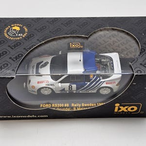 Op de afbeelding: Een model van een Ford RS200 #8 Rally Sweden 1986 auto in een displaydoos. De witte auto heeft blauwe en zwarte accenten, het nummer 8 en Pirelli-branding. De zwarte doos heeft het "ixo models" logo en tekst.