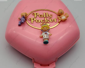 Może przedstawiać: Różowa plastikowa zabawka Polly Pocket z złotym logo w kształcie serca, na którym widnieje napis "Polly Pocket". Zabawka ma klapkę i zawiera trzy małe plastikowe figurki: dziecko, lekarza i pielęgniarkę.