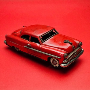 Pode incluir: Um carro de brinquedo vermelho vintage com pneus de parede branca e detalhes cromados. O modelo clássico é colocado sobre um fundo vermelho sólido, mostrando seu design detalhado e aparência envelhecida. Um item de colecionador nostálgico.