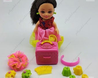May include: A pink Polly Pocket playset with a doll, a pink suitcase, and various accessories. The accessories include a yellow and green headband, a yellow and green hair comb, a green and yellow flower hair clip, a green and yellow flower headband, and a pink flower headband.