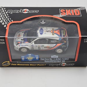 Op de afbeelding: Een Ford Focus WRC modelauto in een zwarte doos. De witte auto heeft blauwe en oranje accenten, het nummer 5 en Martini-branding. De doos is gemarkeerd met "Limited Edition" en "Rally Winners Collection".