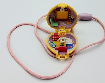 Può includere: Un ciondolo a forma di cuore giallo con un cordino rosa. Il ciondolo è aperto e rivela una bambola in miniatura all'interno con un letto, una cassettiera e uno specchio.