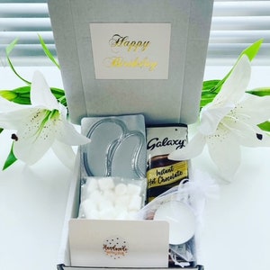 May include: A white gift box with a gold inscription that says "Happy Birthday". The box contains a variety of items, including a silver eye mask, a bar of Galaxy chocolate, a packet of instant hot chocolate, a bag of marshmallows, and a small candle.