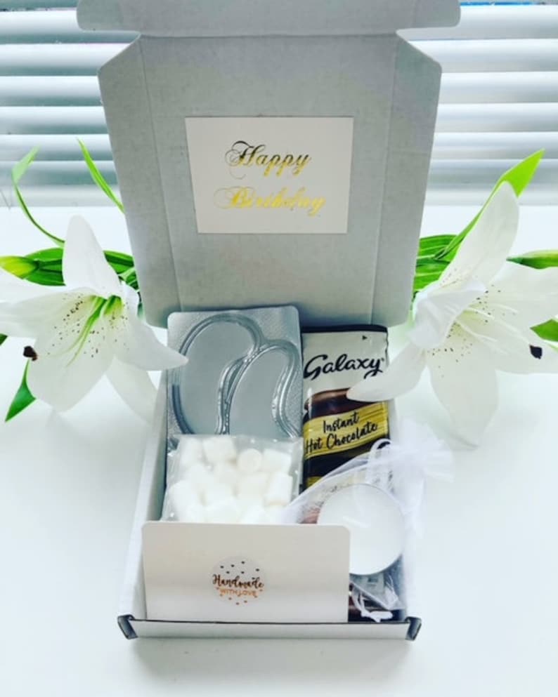 May include: A white gift box with a gold inscription that says "Happy Birthday". The box contains a variety of items, including a silver eye mask, a bar of Galaxy chocolate, a packet of instant hot chocolate, a bag of marshmallows, and a small candle.