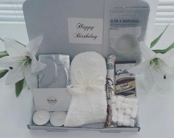 May include: A white gift box filled with a variety of self-care items, including a pair of eye masks, a white fuzzy sock, a chocolate bar, a packet of hot chocolate mix, and marshmallows. The box also includes two white candles and a card that says "Happy Birthday".