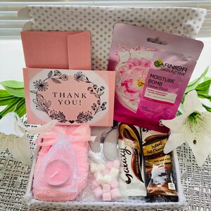 May include: A gift box filled with self-care items, including a pink face mask, a pink candle, a pair of pink fluffy socks, a packet of marshmallows, a hot chocolate packet, and a thank you card.