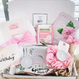 May include: A gift box containing self-care products. It features a pink scrunchie, eye masks, a Galaxy Ripple chocolate bar, a tea bag, a face mask, a candle, and a card that says "Happy Birthday".