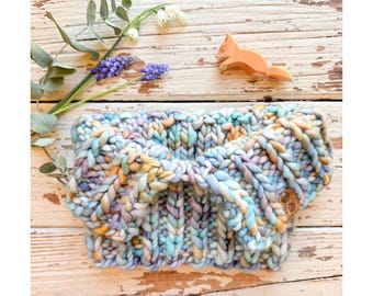 May include: A chunky knit headband in pastel colors of blue, yellow, and purple. The headband is made of thick yarn and features a knotted design. A small wooden fox and sprigs of lavender and white flowers are in the background.