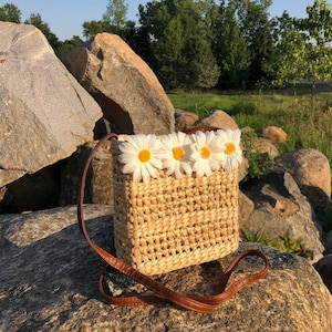 May include: A woven straw bag with a brown leather strap and a row of white daisy flowers across the top.
