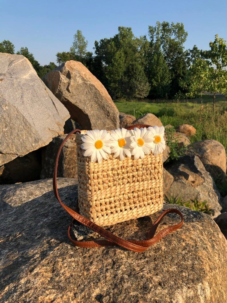 May include: A woven straw bag with a brown leather strap and a row of white daisy flowers across the top.