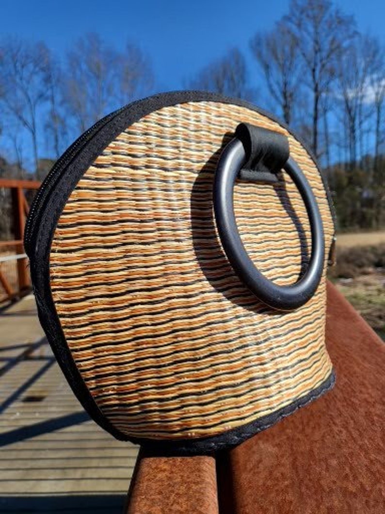Peut inclure: Un sac &agrave; main en paille tress&eacute;e avec une poign&eacute;e en cuir noir. Le sac a une forme arrondie et une bordure noire.