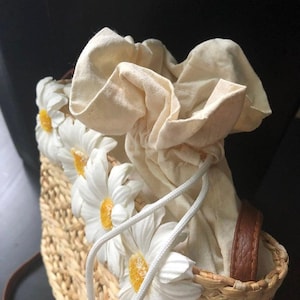 May include: A woven straw bag with a white cotton drawstring closure and white daisy embellishments.