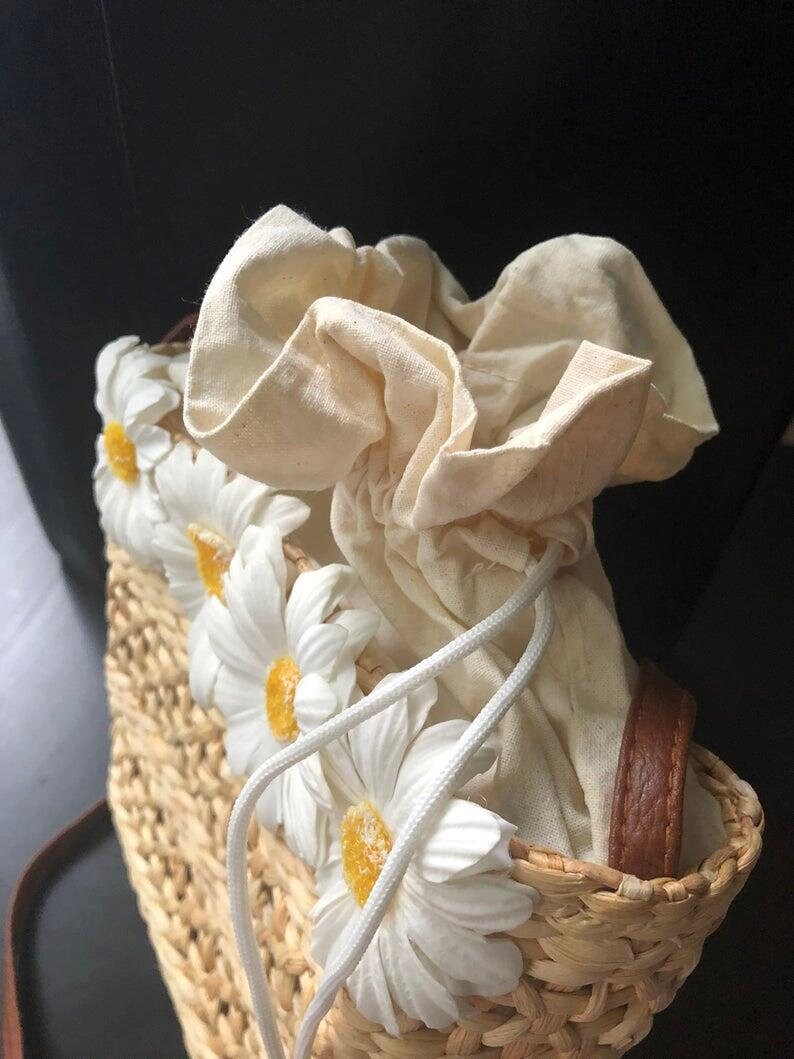May include: A woven straw bag with a white cotton drawstring closure and white daisy embellishments.