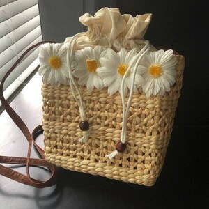 May include: A woven straw bag with a brown leather strap and white daisy embellishments.