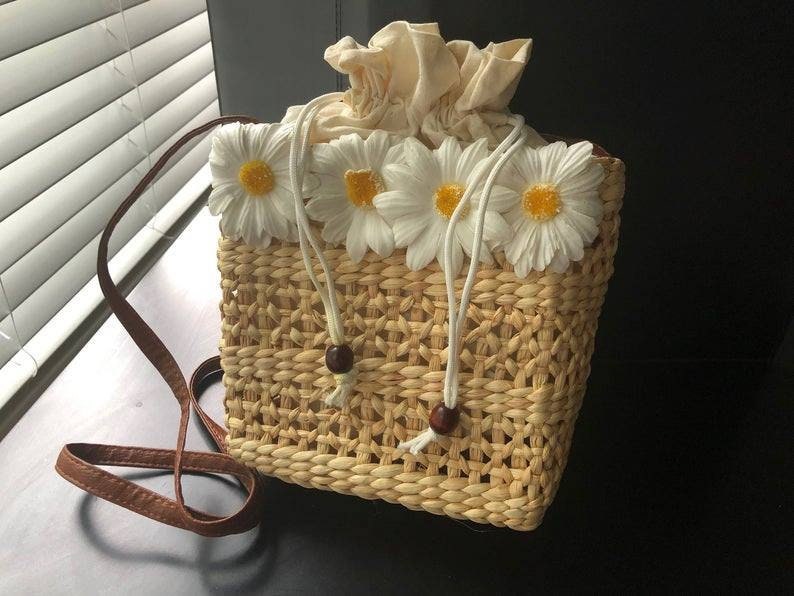 May include: A woven straw bag with a brown leather strap and white daisy embellishments.
