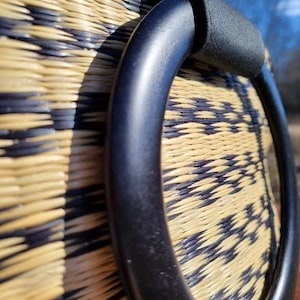 Peut inclure: Gros plan sur une poign&eacute;e tiss&eacute;e noire sur un sac en paille. La poign&eacute;e est faite d'un mat&eacute;riau noir &eacute;pais et a une surface lisse et arrondie.